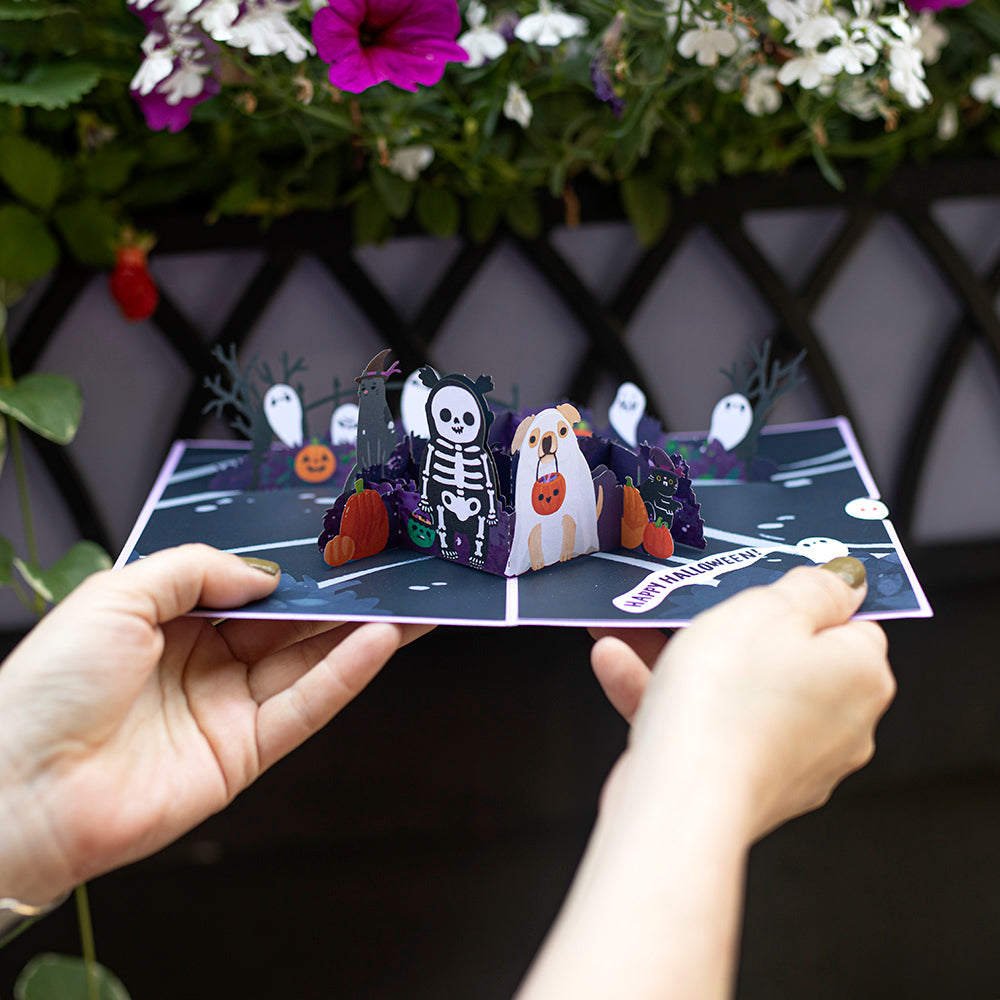 Hands holding an open Halloween pop-up card with paper ghosts, a skeleton and pumpkins